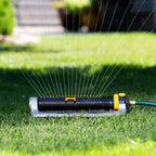 An oscillating lawn sprinkler is connected to a green garden hose, spraying water across a green lawn, with a blurred house in the background.