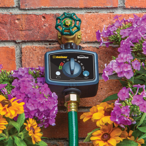 An automatic water timer is attached to a brass spigot on a brick wall, with a green hose connected below it, surrounded by colorful flowers.