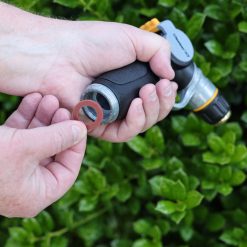 A person's hands holding a garden hose nozzle and a washer, with a backdrop of green foliage. The nozzle appears to be disassembled for maintenance or repair.