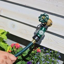 A person is holding a green garden hose with a nozzle attached to a brass outdoor tap against a white wooden background, surrounded by colorful flowers.