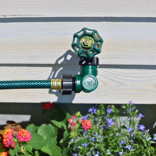 An outdoor water faucet with a green hose attached is mounted on a wooden siding wall, surrounded by colorful flowers in sunlight.