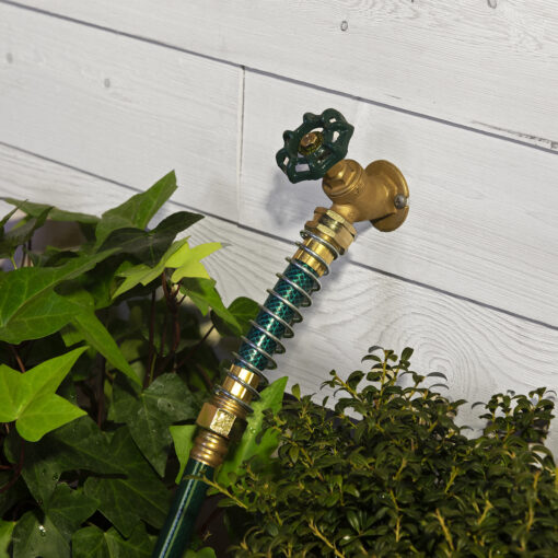 A brass outdoor water tap is attached to a white wooden wall, with a green hose connected to it. Lush green plants surround the bottom area.