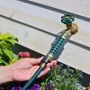 A person's hand is holding a green hose connected to a brass garden tap with an ornate handle, surrounded by sunlit wooden panels and blooming flowers.