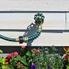 A green garden hose with a brass nozzle is connected to an outdoor spigot on a white wooden wall, surrounded by colorful flowers in sunlight.