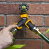 A person's hand is attaching a green garden hose to a yellow and black splitter connected to a brass outdoor faucet against a brick wall.