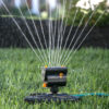 An oscillating sprinkler is watering a lush green lawn, with water droplets creating a sparkling effect in the sunlight, against a blurred garden backdrop.