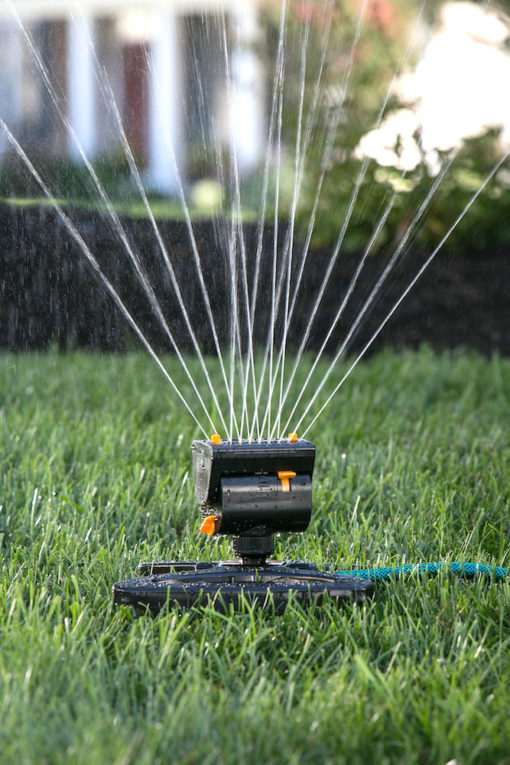 An oscillating sprinkler is watering a lush green lawn, with water droplets creating a sparkling effect in the sunlight, against a blurred garden backdrop.