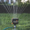 A stationary oscillating lawn sprinkler is watering fresh green grass, with a gentle mist of water droplets captured in sunlight against a soft-focus background.