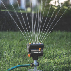 A stationary oscillating lawn sprinkler is watering fresh green grass, with a gentle mist of water droplets captured in sunlight against a soft-focus background.