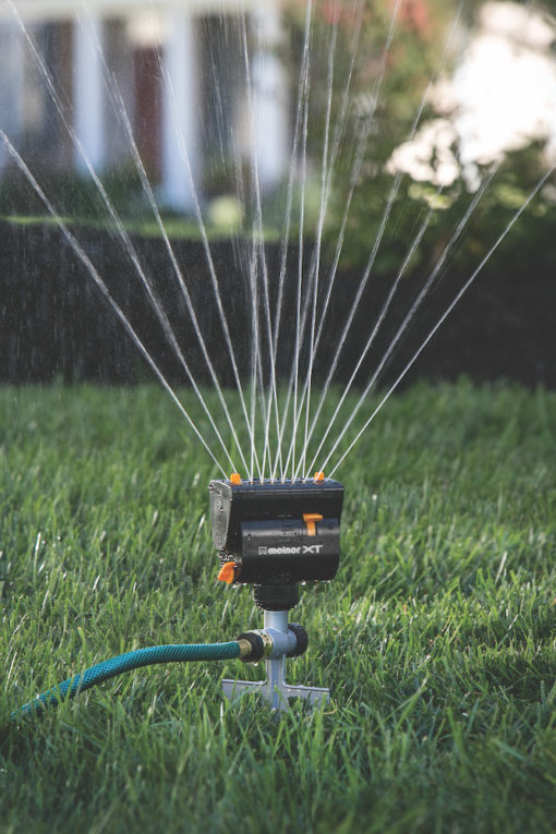 A stationary oscillating lawn sprinkler is watering fresh green grass, with a gentle mist of water droplets captured in sunlight against a soft-focus background.