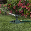 This image shows a green lawn sprinkler in action on a vibrant grassy area, with water spraying out amidst a backdrop of pink and red flowers.