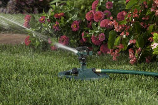 This image shows a green lawn sprinkler in action on a vibrant grassy area, with water spraying out amidst a backdrop of pink and red flowers.