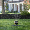 An oscillating sprinkler is watering a lush green lawn in a residential area with a house visible in the background on a sunny day.