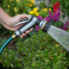 A person's hand holds a hose with a spray nozzle, watering green plants and flowers in a garden, showcasing a gardening activity.