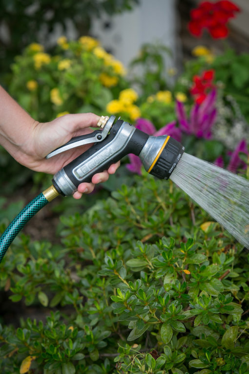 A person's hand holds a hose with a spray nozzle, watering green plants and flowers in a garden, showcasing a gardening activity.