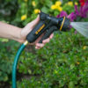 A person's hand is holding a garden hose with a nozzle, spraying water onto green plants, with bright flowers in the background.