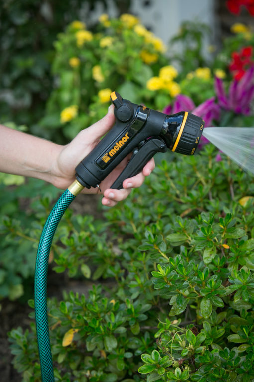 A person's hand is holding a garden hose with a nozzle, spraying water onto green plants, with bright flowers in the background.