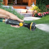 A hand holding the Melnor RelaxGrip straight hose nozzle spraying water with the full spray pattern against a backdrop of green grass leading to a stone walkway and wooden deck.