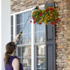 A person is watering a colorful hanging flower basket beside a window on a house with stone wall accents, using a long reach hose nozzle.