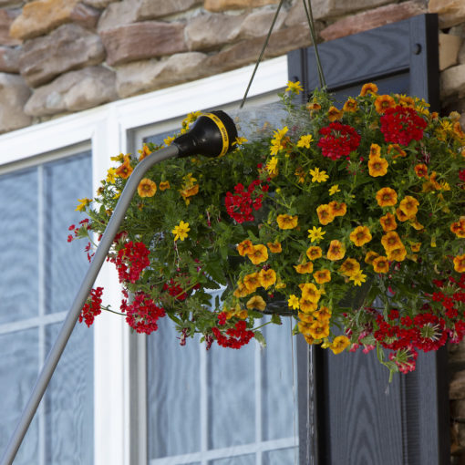 A lush hanging basket filled with red and yellow flowers is watered by a long white hose with a black nozzle against a stonewall backdrop.