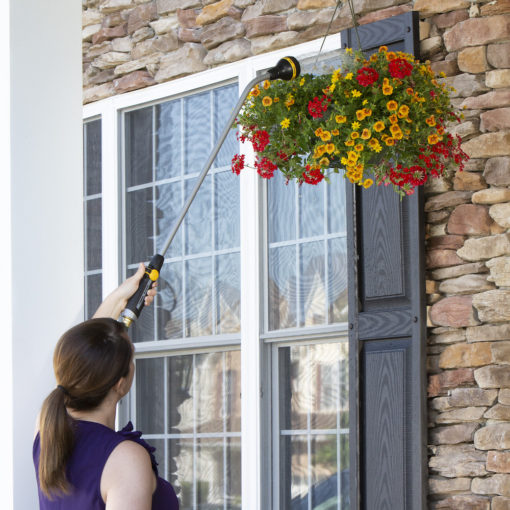 A person is watering a hanging basket of vibrant red and yellow flowers outside a stone-faced house with a black door and white-framed windows.