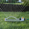 An oscillating lawn sprinkler is connected to a green hose, spraying water in an arch pattern over a well-manicured grassy lawn with a mulched area behind.