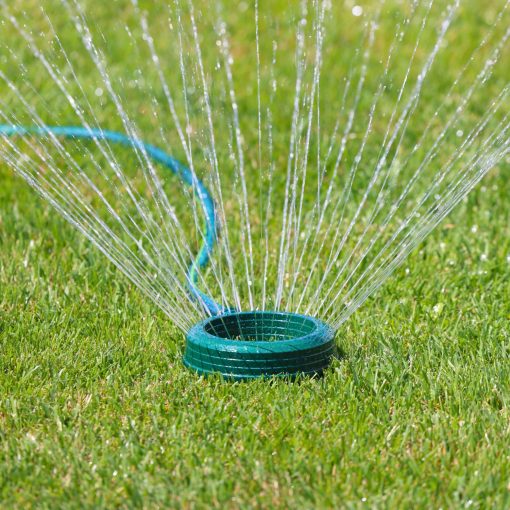 A circular lawn sprinkler is actively watering the grass, creating a radial pattern of water jets against a background of vibrant green lawn.