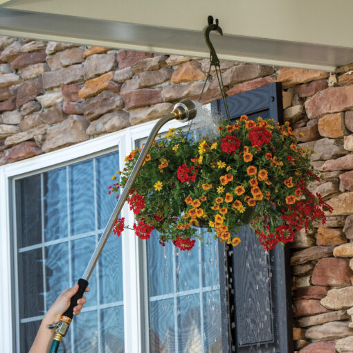 A person is watering a lush, colorful hanging basket of flowers using an extendable watering wand outside a stone-facade house with windows.