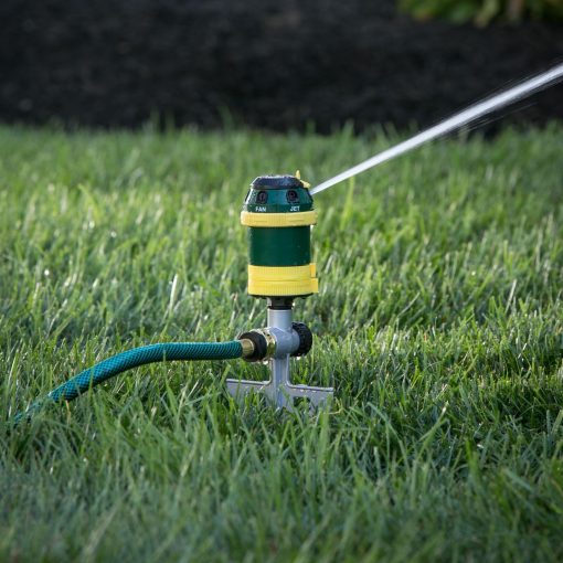 A sprinkler is attached to a green hose, spraying water onto lush grass. The background is slightly shaded, indicating it might be early morning or late afternoon.