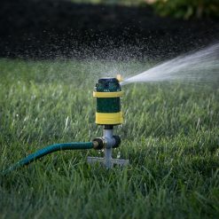 A green and yellow sprinkler is connected to a hose, spraying water over a grassy lawn with sunlight casting a warm glow in the background.