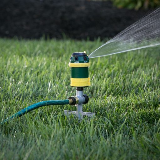 A garden sprinkler attached to a hose is actively watering the lawn, spraying a fine mist over green grass with dark soil in the background.