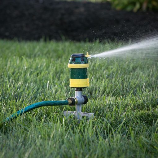 A green and yellow sprinkler attached to a hose is actively watering a lush lawn with a fine mist, set against a backdrop of dark mulch.