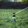 A green and yellow garden sprinkler is connected to a hose, actively spraying water on a well-manicured, vibrant green lawn with a dark mulch background.