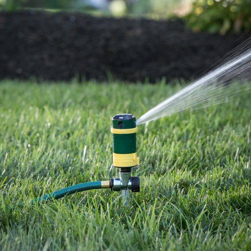 A garden sprinkler is attached to a green hose, actively spraying water over lush grass with a dark mulch bed in the background.