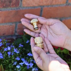 A person is holding two brass hose connectors in front of a brick wall with blue flowers in the background. It's a sunny day.