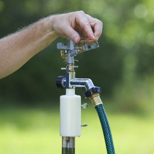 A person's hand turns a water spigot with a hose attached. The scene is outdoors, with a blurry green background suggesting a garden setting.