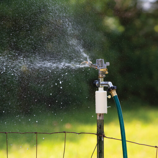 This image shows a sprinkler attached to a garden hose, spraying water onto a lawn. The mechanism is fixed to a wire fence, with greenery in the background.