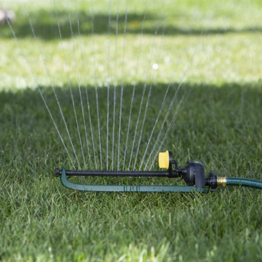 An oscillating sprinkler attached to a green hose is watering the lawn, casting a spray pattern on a bright, sunny day with green grass.