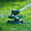 An oscillating lawn sprinkler is attached to a hose, actively watering the surrounding green grass on a sunny day. It's highlighted by sunlight and shadows.