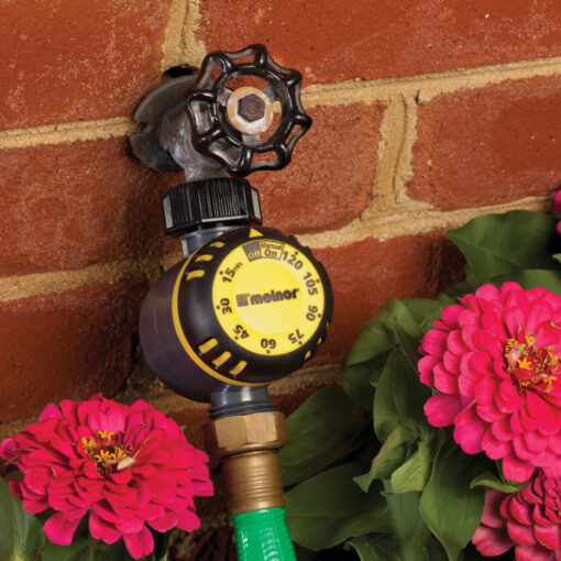 An outdoor water faucet with a timer attached is installed on a red brick wall, alongside blooming pink flowers, possibly in a garden.