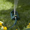 In the image, there is an oscillating lawn sprinkler watering grass, surrounded by green foliage and yellow flowers, under bright sunlight.