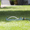 An oscillating sprinkler is attached to a garden hose, actively spraying water over a well-manicured green lawn with a blurred background suggesting a residential garden setting.