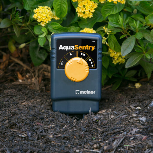 An AquaSentry moisture sensor by Melnor is placed in soil among flowering plants. It appears to be a garden watering automation tool.