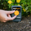 A person's hand is adjusting a yellow dial on a Melnor AquaSentry moisture sensor, which is placed in soil near green shrubbery.