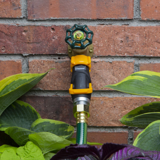 A garden hose nozzle attached to a green hose is set against a red brick wall, surrounded by lush green plant leaves.