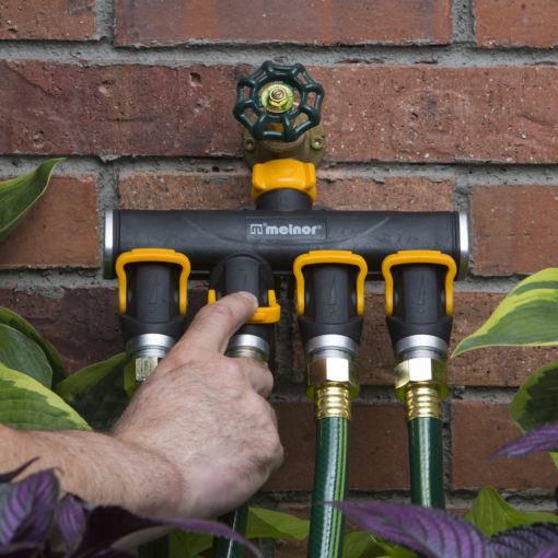 A person's hand is adjusting a Melnor four-way hose distributor attached to a brick wall, with connected green hoses and surrounded by plants.