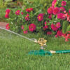 An oscillating lawn sprinkler is attached to a green hose, actively watering a vibrant green lawn with a backdrop of colorful pink and red flowers.