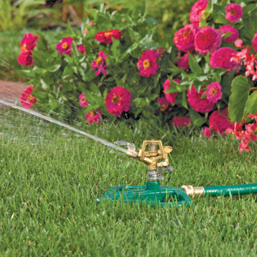 An oscillating lawn sprinkler is attached to a green hose, actively watering a vibrant green lawn with a backdrop of colorful pink and red flowers.