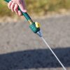 A person's hand is holding a green garden hose with a yellow lever, spraying water onto a dry, cracked ground. It's a close-up shot in daylight.