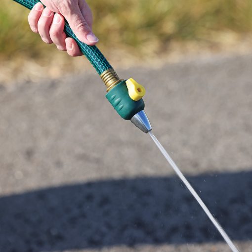 A person's hand is holding a green garden hose with a yellow lever, spraying water onto a dry, cracked ground. It's a close-up shot in daylight.